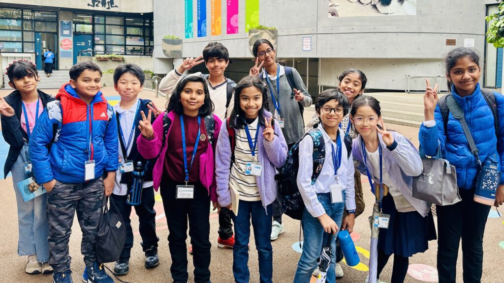 Group of smiling students posing outdoors, flashing peace signs, wearing backpacks and ID badges at a school entrance.