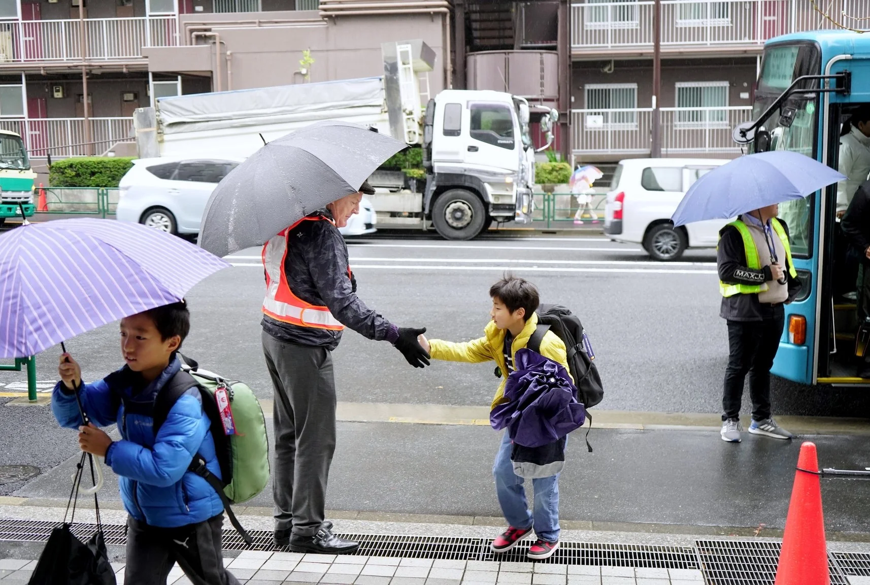 Children with umbrellas board a bus in rainy weather. Helper in vest assists and shakes hands with a child.