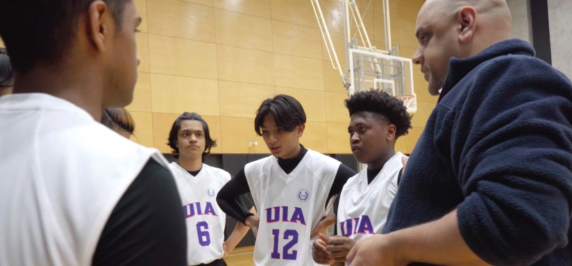 Basketball coach gives strategy talk to players in white jerseys on indoor court.