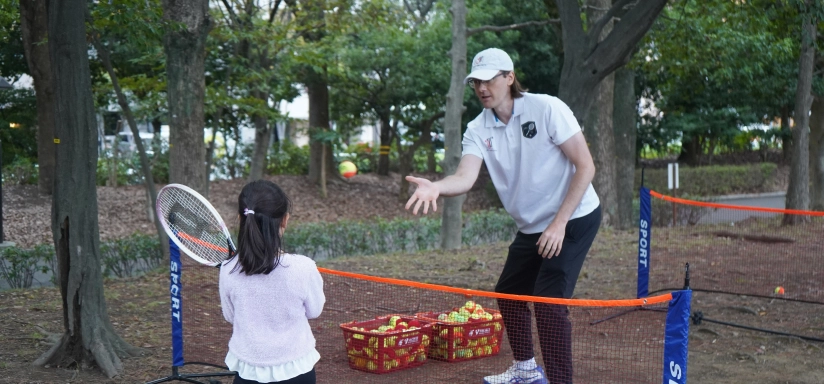 Tennis coach teaching a young girl to play outdoors with a mini net and rackets in a park setting.