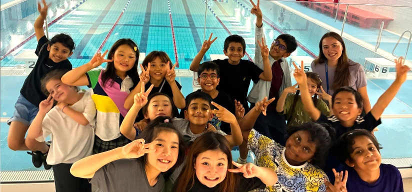 Group of smiling kids and a teacher posing with peace signs by a swimming pool indoors.