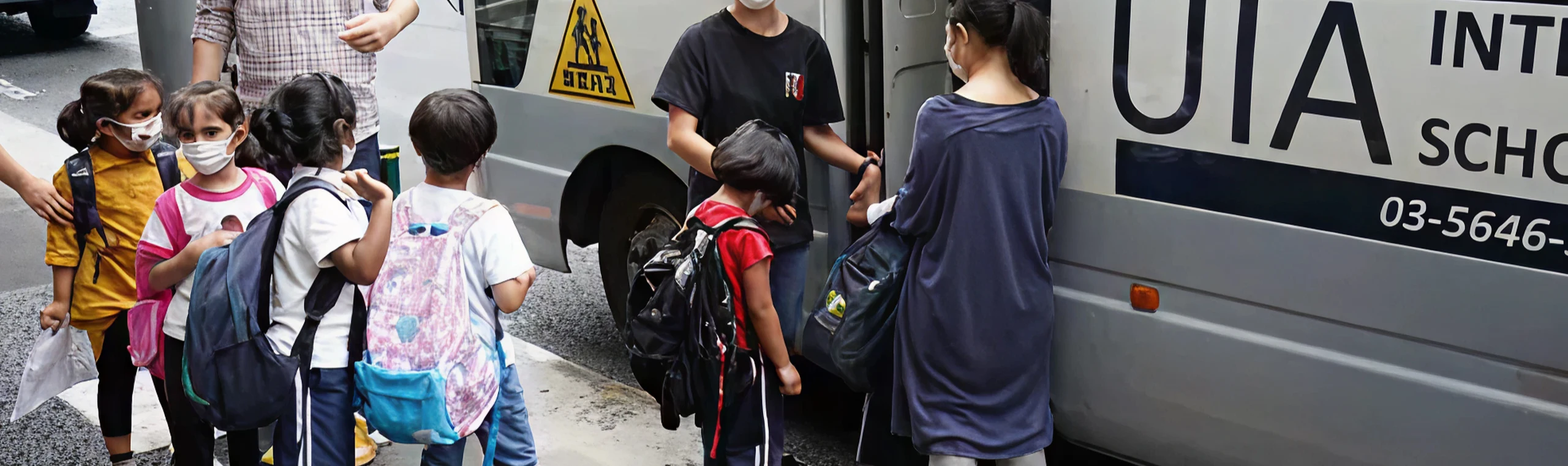 Children boarding a school bus with backpacks, some wearing masks, guided by adults on a city street.
