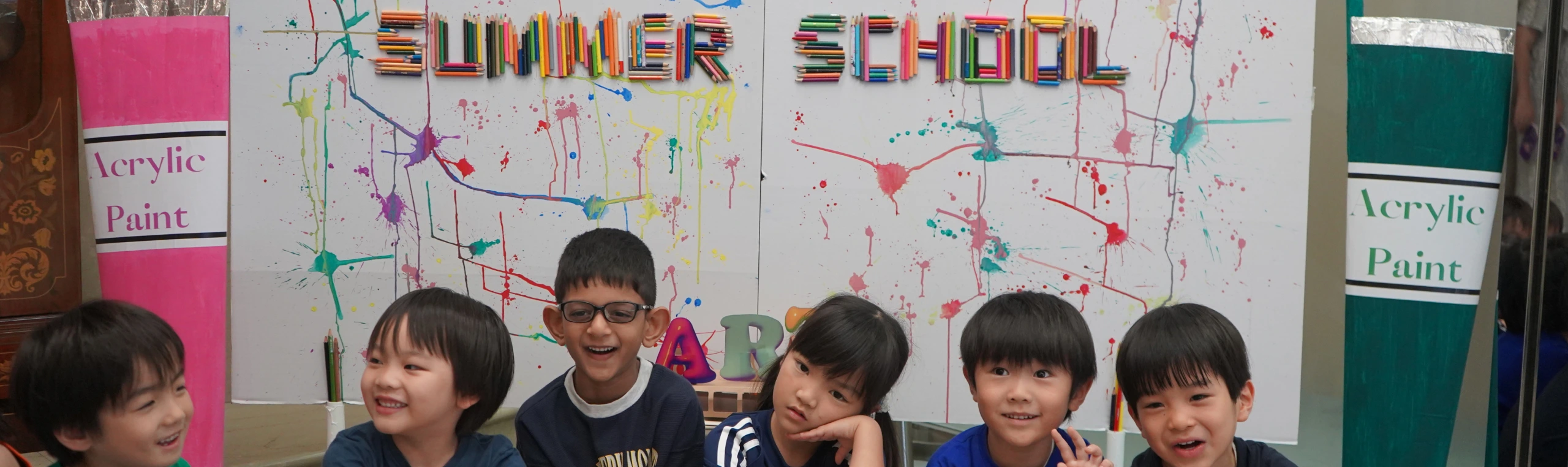 Five children smiling in front of a colorful Summer School art display with paint splatters.