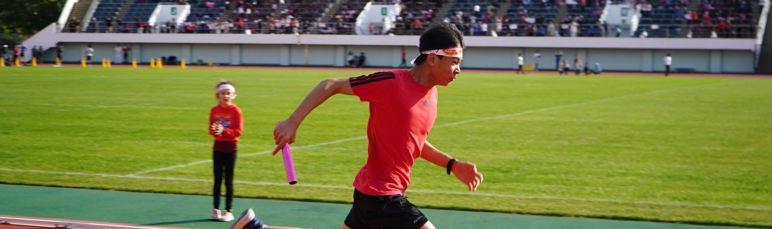 Athlete in red running relay race with baton on sunny day at stadium, spectators in background.
