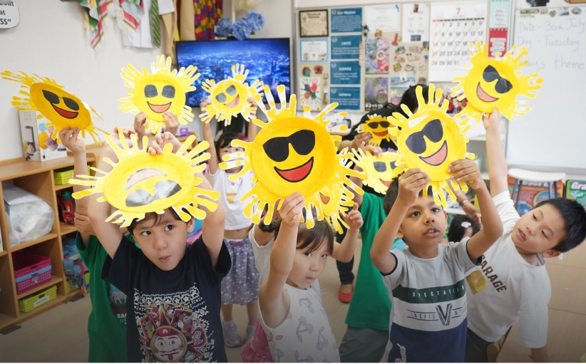 Children holding sun-shaped crafts with sunglasses, smiling in a colorful classroom.