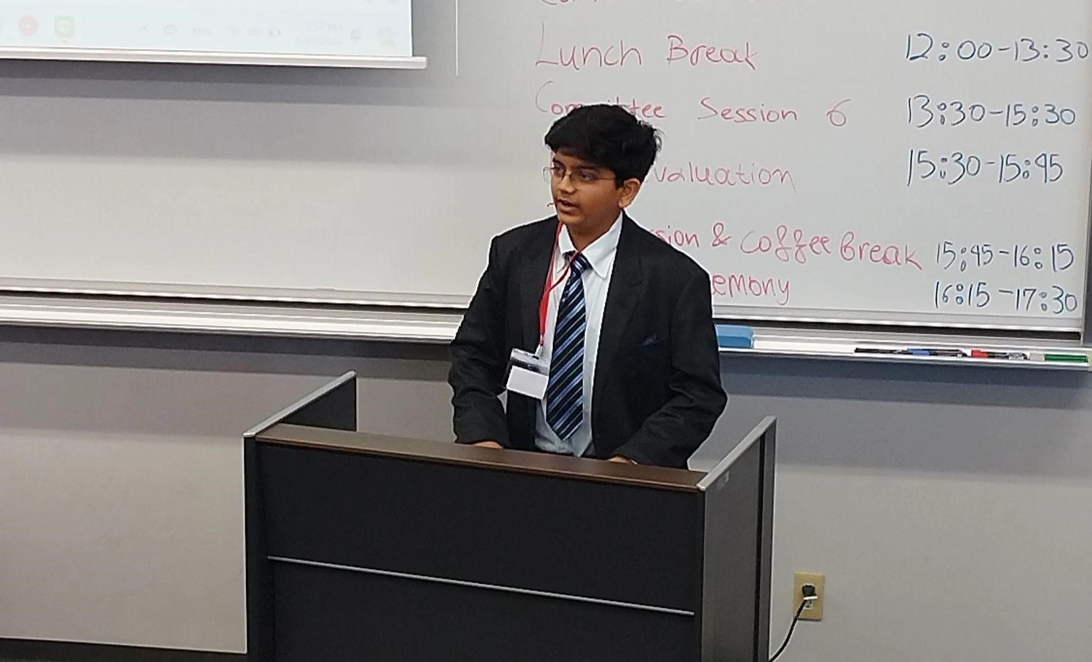 Young speaker presenting at a conference podium with a schedule on a whiteboard behind him.