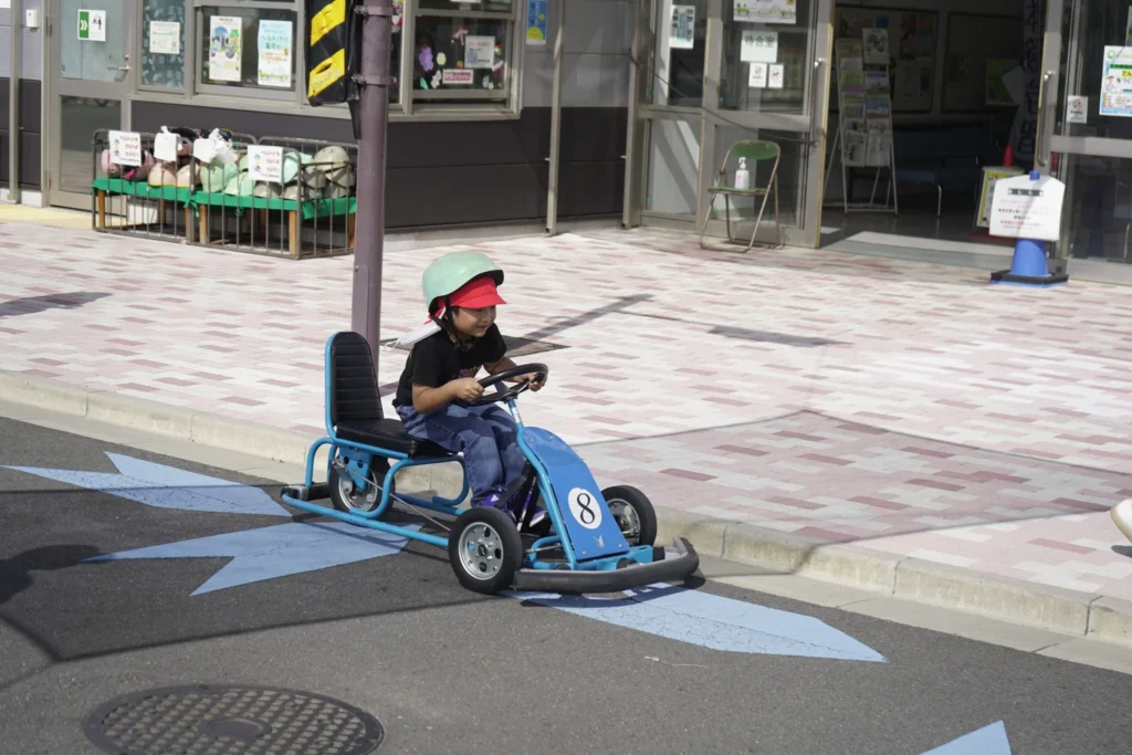 Child driving a blue pedal kart on a sunny day, wearing a helmet and cap, outdoors near a building.
