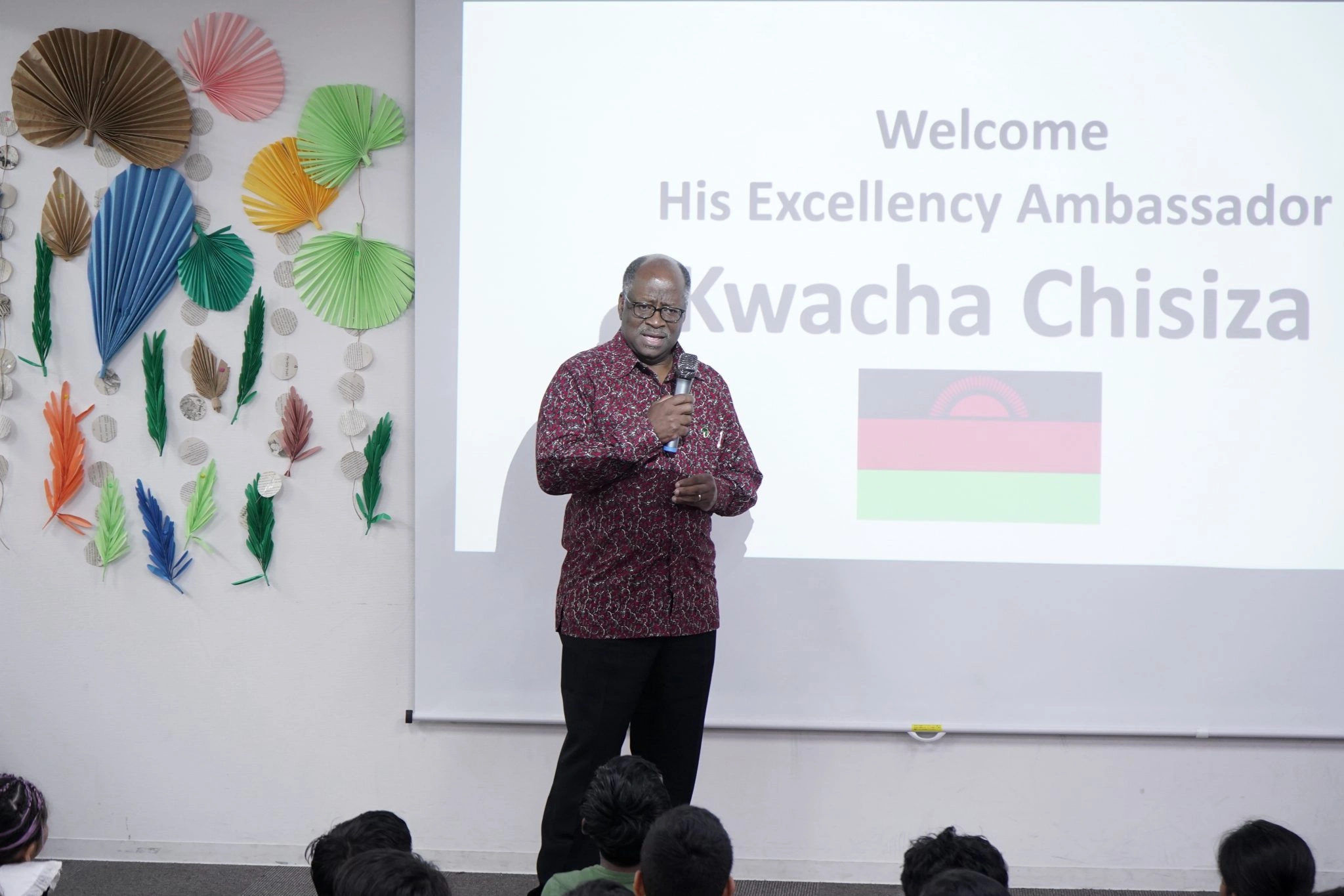 Man speaking at an event with colorful decorations, projected text with Welcome His Excellency Ambassador, flag in view.