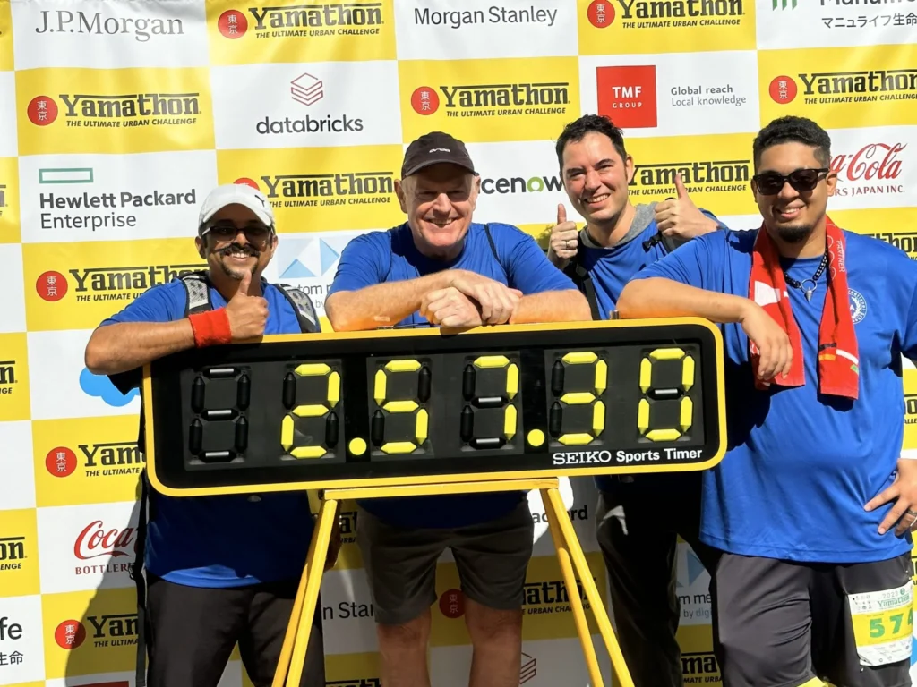 Four smiling participants pose with a sports timer at YamaTHON 2023 against a backdrop of sponsor logos.