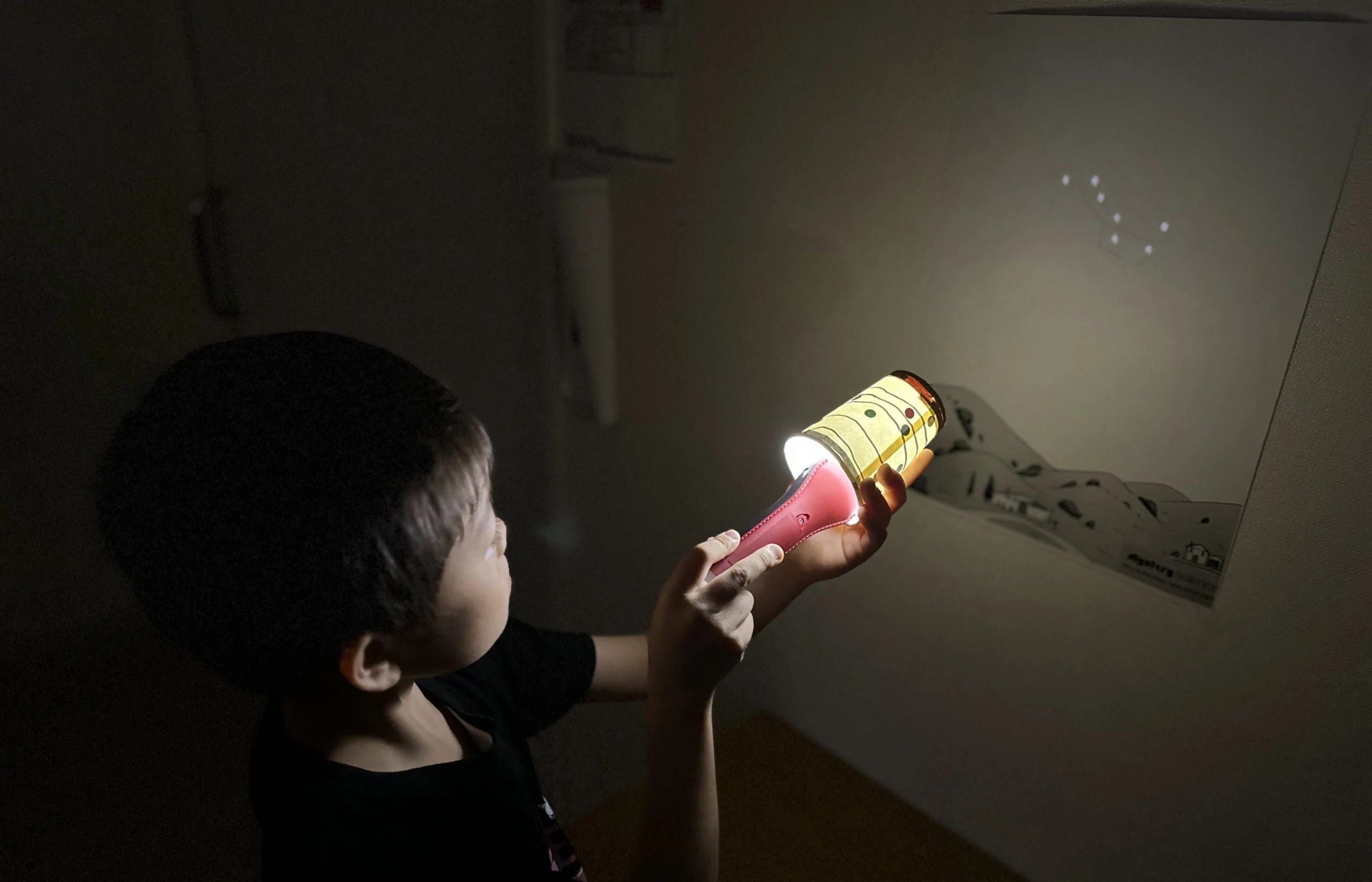 Child using a flashlight to project stars on the wall, learning about constellations in a dark room.