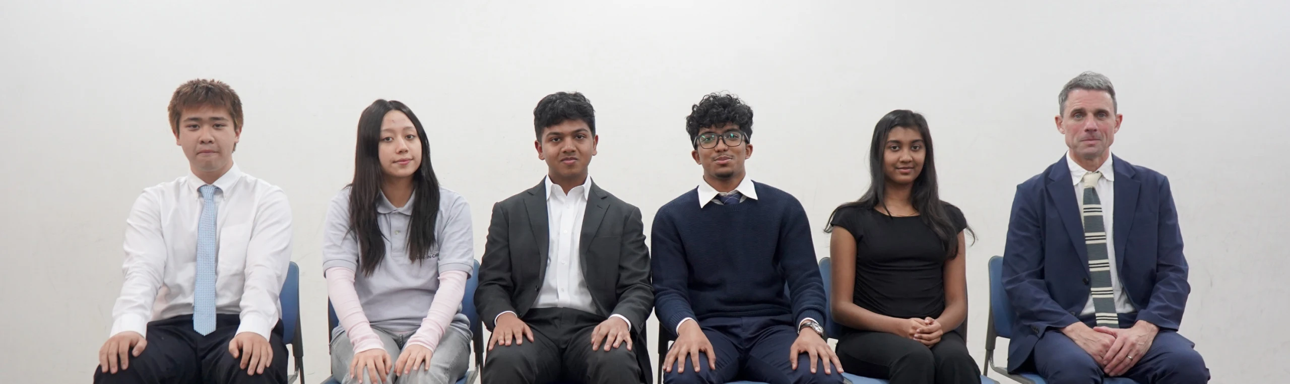 A diverse group of six people sitting in a row, dressed in business attire against a plain background.