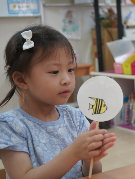 Child holding a handmade fish art craft in a classroom, focusing intently.