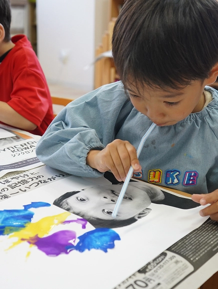 Child painting with straw, creating colorful abstract art on paper. Engaged in creative activity at a classroom table.