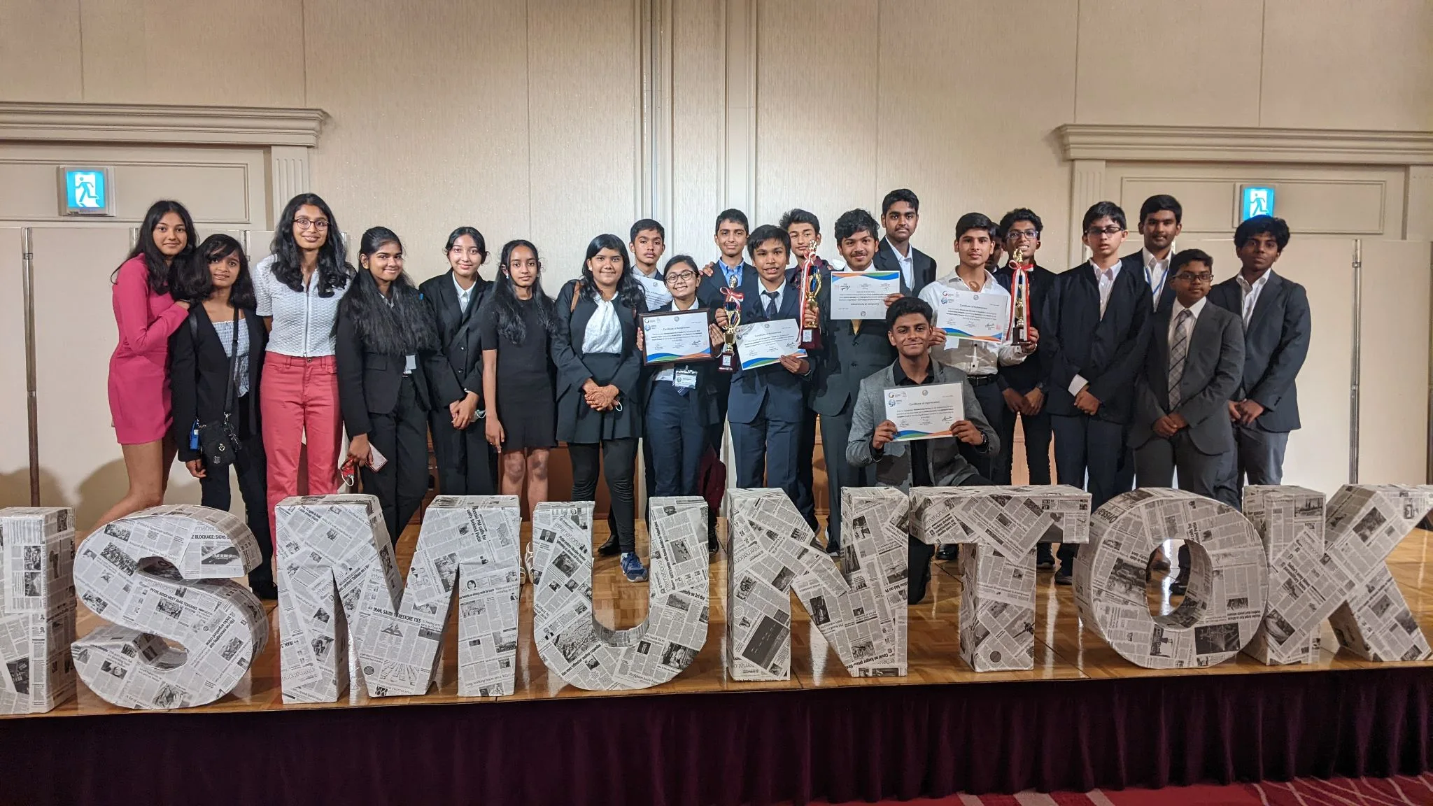 Group of students in formal attire holding certificates and trophies at an event, standing behind large ISMUN TOK letters.