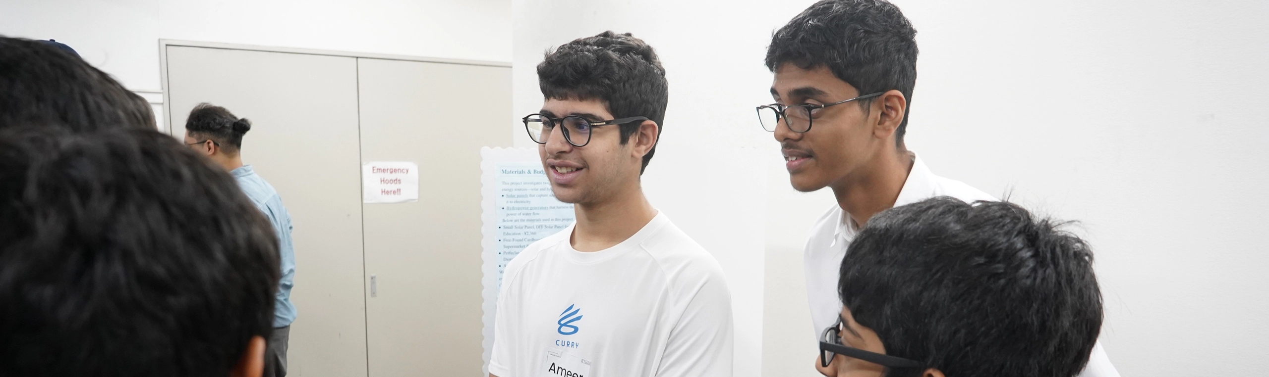 Group of young students engaged in discussion at a science exhibition, wearing white shirts and glasses.