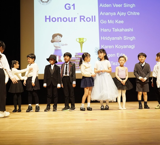 Children standing on stage for G1 Honour Roll ceremony, names displayed on screen in the background.
