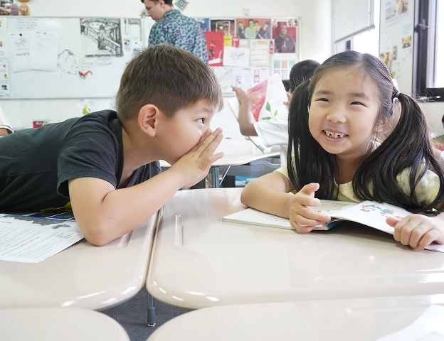 Two children smiling and whispering in a classroom setting, with books on their desks.