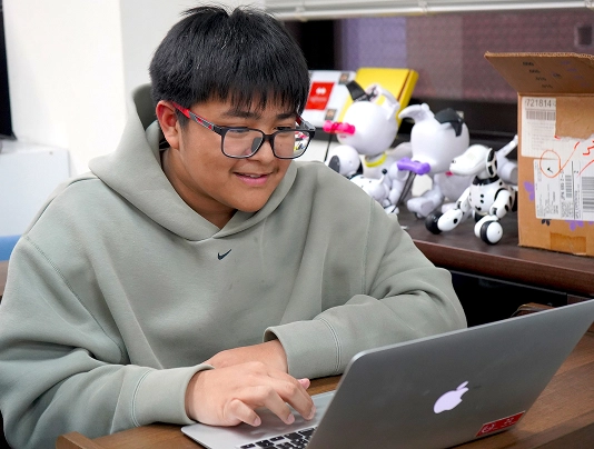 Person in gray hoodie using laptop, surrounded by robotic toys and boxes on a desk, focused and engaged in technology.