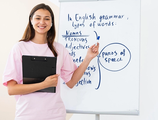 Smiling woman teaching English grammar, points to parts of speech on a whiteboard, holding a clipboard.