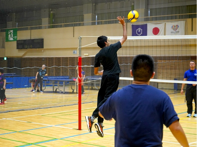 Athlete spikes volleyball during indoor match, while teammates and opponents watch in a gym setting.