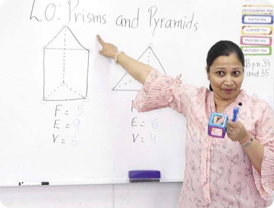 Teacher explaining prisms and pyramids with diagrams on a whiteboard in a classroom setting.