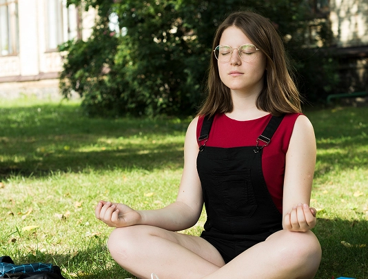 Person meditating outdoors, sitting cross-legged on grass, wearing red and black, surrounded by greenery.