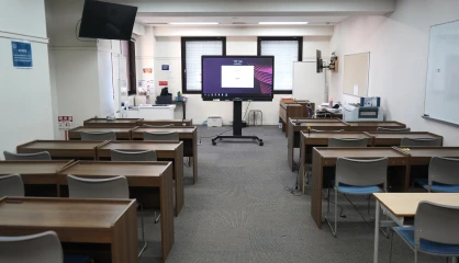 Empty classroom with desks, chairs, a digital display board, and a whiteboard, ready for a lecture or presentation.