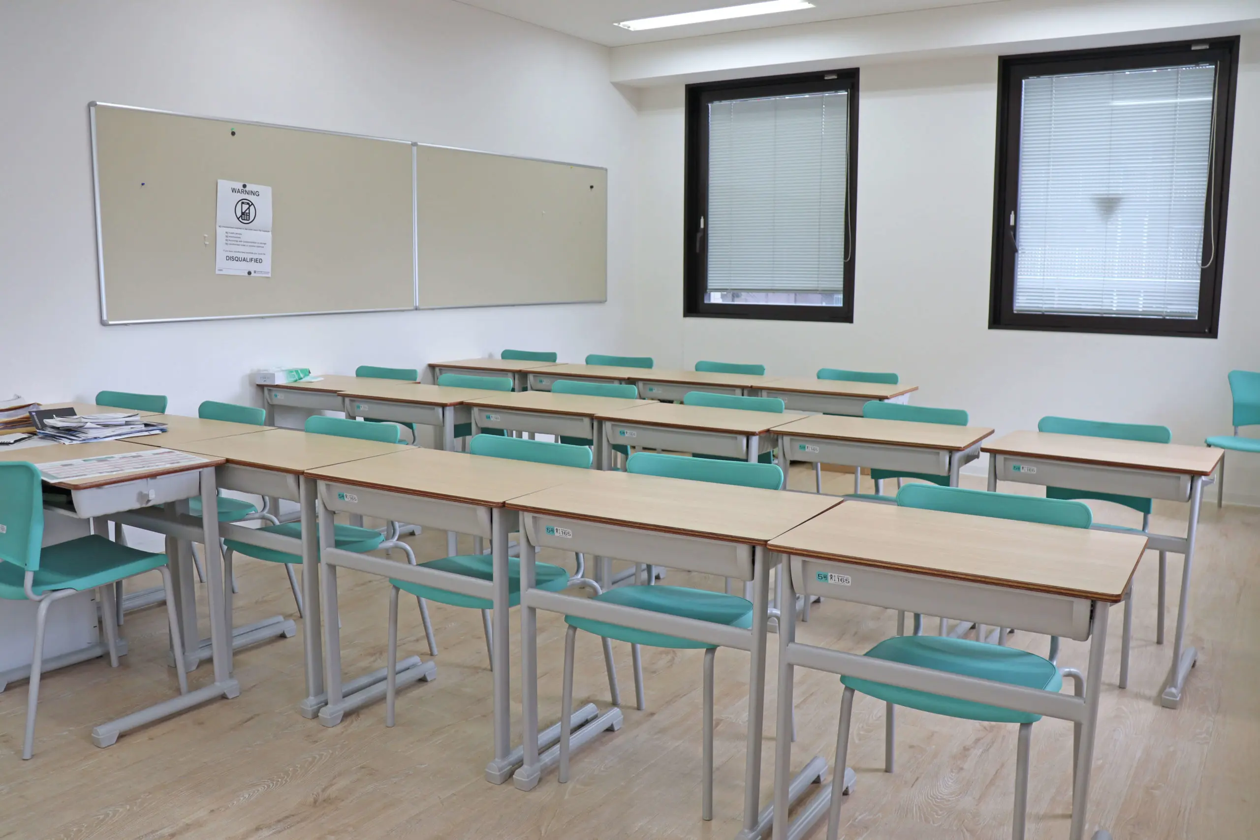 Empty classroom with wooden desks and teal chairs, whiteboard, and windows with blinds. Bright and organized learning space.